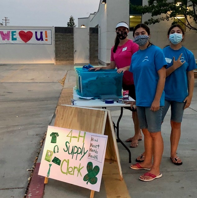 3 youth with face masks standing at table with 4-H supply clerk sign