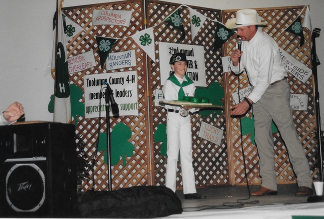 Maddalena as a young girl wearing her 4-H uniform as a man in a cowboy gestures towards her and speaks into a microphone.