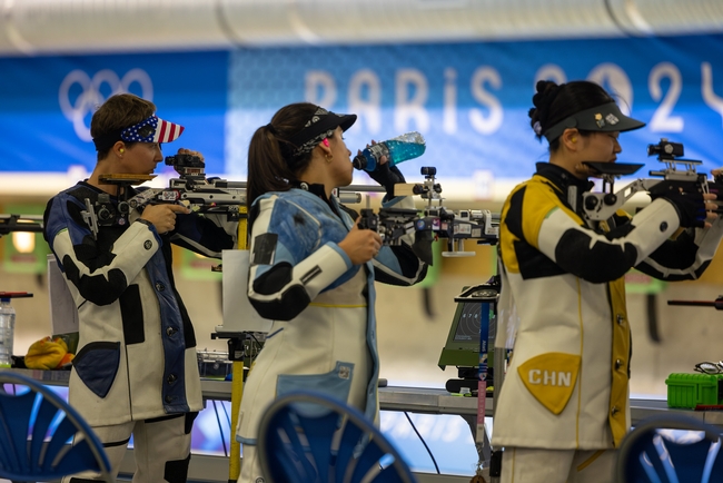 Maddalena, wearing the U.S. Olympic uniform and a visor with a flag design, stands beside two Olympic competitors and their rifles.