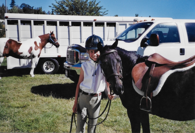 Shown as a youngster, Maddalena holds the reins of her horse. A horse and white horse trailer in the background.