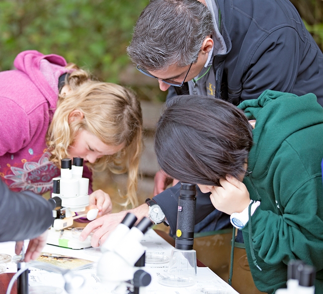 Lacan adjusts a petri dish under a microscope as two girls look into microscopes.