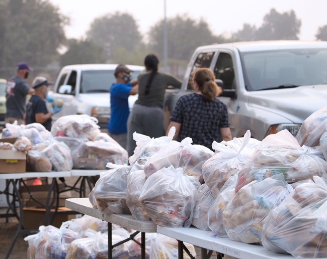 In the foreground are tables with mounds of white plastic bags filled with food items. In the background are volunteers wearing facemasks placing bags of food in the back of a silver pickup and a white pickup.