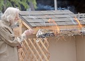 Steve Quarles gestures toward flames rising from dry pine needles and leafy debris in a melting plastic rain gutter affixed to a structure with a shingled roof.