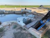 Gates are open, releasing water into an irrigation ditch running between a road and a crop field.