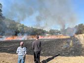 Two people stand on the edge of a blackened field with a line of flames.