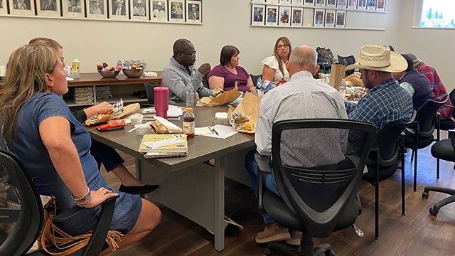 Nine people - men and women - sit around a conference room table.