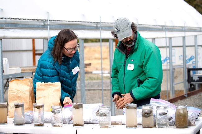 A woman in a puffy blue jacket and a bearded man in a green jacket look at slake test results in glass jars.