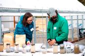 A woman in a puffy blue jacket and a bearded man in a green jacket look at slake test results in glass jars.