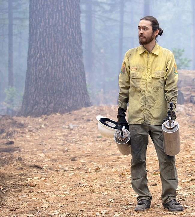 Wearing a yellow shirt smudged with soot, Kane holds a drip torch in each hand. He is shown standing on a forest floor littered with dry leaves and other vegetation.
