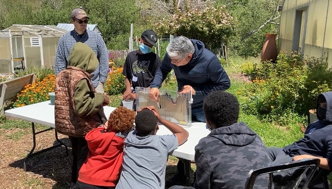 Children gather around a table watching as Sandoval points to the water flowing through the sand in the model.