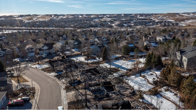 A patch of blackened ruins of houses surrounded by homes with yards blanketed in white snow.