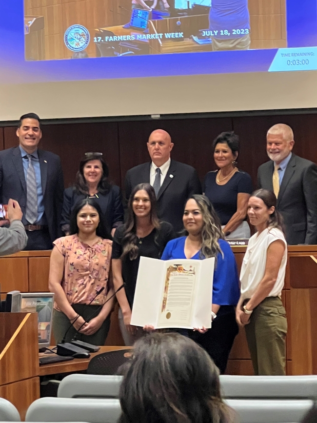 Nine people in two rows posed with a large certificate displayed in the middle. Located in Board chambers
