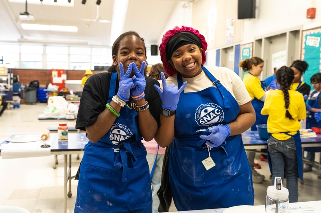 Two girls smiling in a school cafeteria with blue aprons and food prep gloves on.
