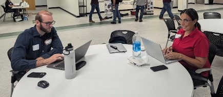 Two people sitting on a table, working on their computers and smiling to the camera.