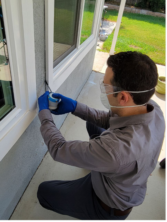 A man wearing a mask, gloves, long sleeve shirt, and pants applying a pesticide dust into a crack on an exterior wall.