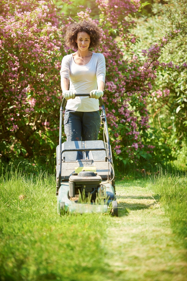 A person with brown curly hair in a white long sleeve shirt pushing a lawnmower through green grass.