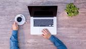 An overhead shot of a person with one hand holding a coffee mug and the other hand on the mousepad of a laptop.