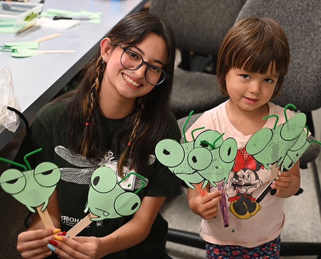 Bohart Museum intern Melody Ruiz, co-coordinator of the Bohart's arts and crafts activities, shows her Mantis on a Stick creations with Maya Lee, 4, of Woodland. (Photo by Kathy Keatley Garvey)