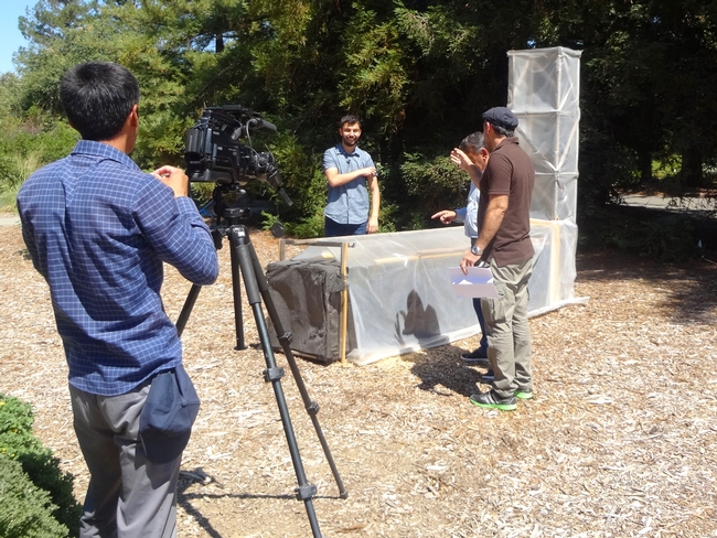 Man with video camera on tripod films three people talking in front of chimney solar dryer at UC Davis.