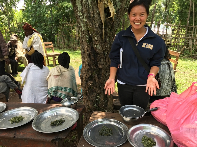 Lauren in UC Davis sweatshirt, pointing to plates of cooked kale and cooked sweet potato leaves, outside in Ethiopia.