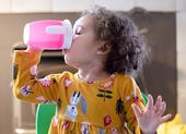 A toddler with curly brown hair drinks water from a pink sippy cup.