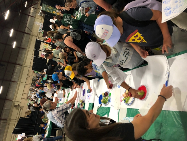 Master Gardeners helping elementary schoolers paint pictures from patterns made from applying paint to celery, apples, and corn. Pictures shows 14 students lined up, each at a painting station with a paint pad and celery, apples, and corn, with Master Gardeners on opposite side of table.