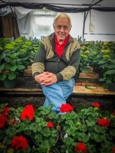 Dr. David Gibby at Gibby Greenhouses - Sitting in greenhouse surrounded by red and yellow flowers.
