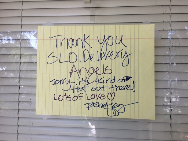 A sign taped on a window with blinds in the background that reads. Thank you SLO Deivery Angels, Sorry-it's kind of hot out there! Lots of Love.