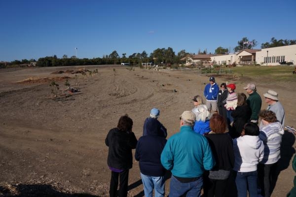 A group of 15 people stand in the corner of a large field with 4 rows. One row is planted with small tree saplings, supported by posts. In the distance, a large white building, the Veterans Home of California – Ventura is visible.