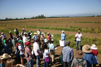 A group of two dozen people in a field listening to a speaker.