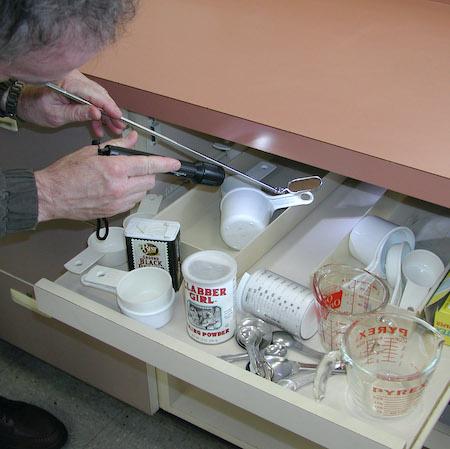 A man uses a small mirror and flashlight to look for cockroaches in kitchen drawers. Credit: Cheryl A. Reynolds, UC IPM.