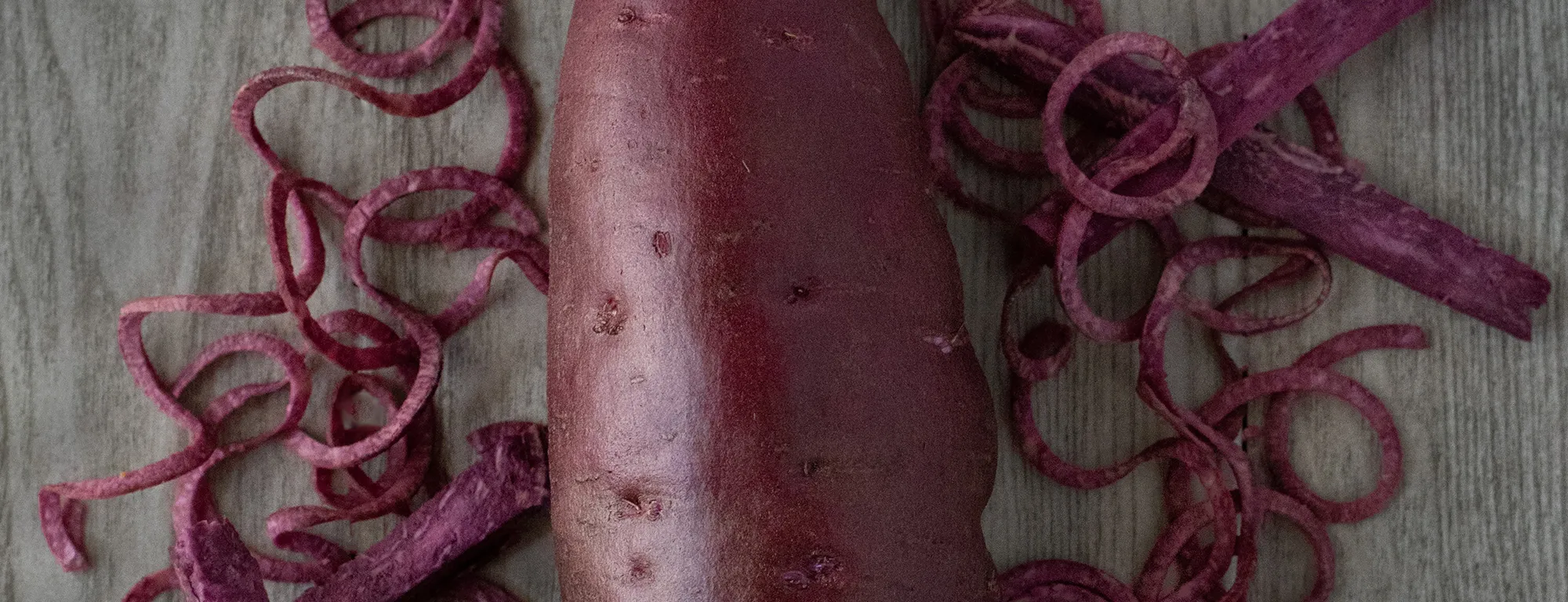 A whole dark purple-skinned sweetpotato flanked by pieces of purple sweetpotato flesh cut into curls