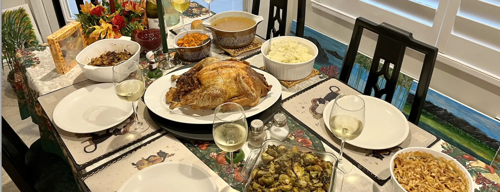 A dining table with stuffing, sweet potatoes, gravy, a roast turkey, mashed potatoes, Brussels sprouts, green bean casserole, and 5 glasses of wine.