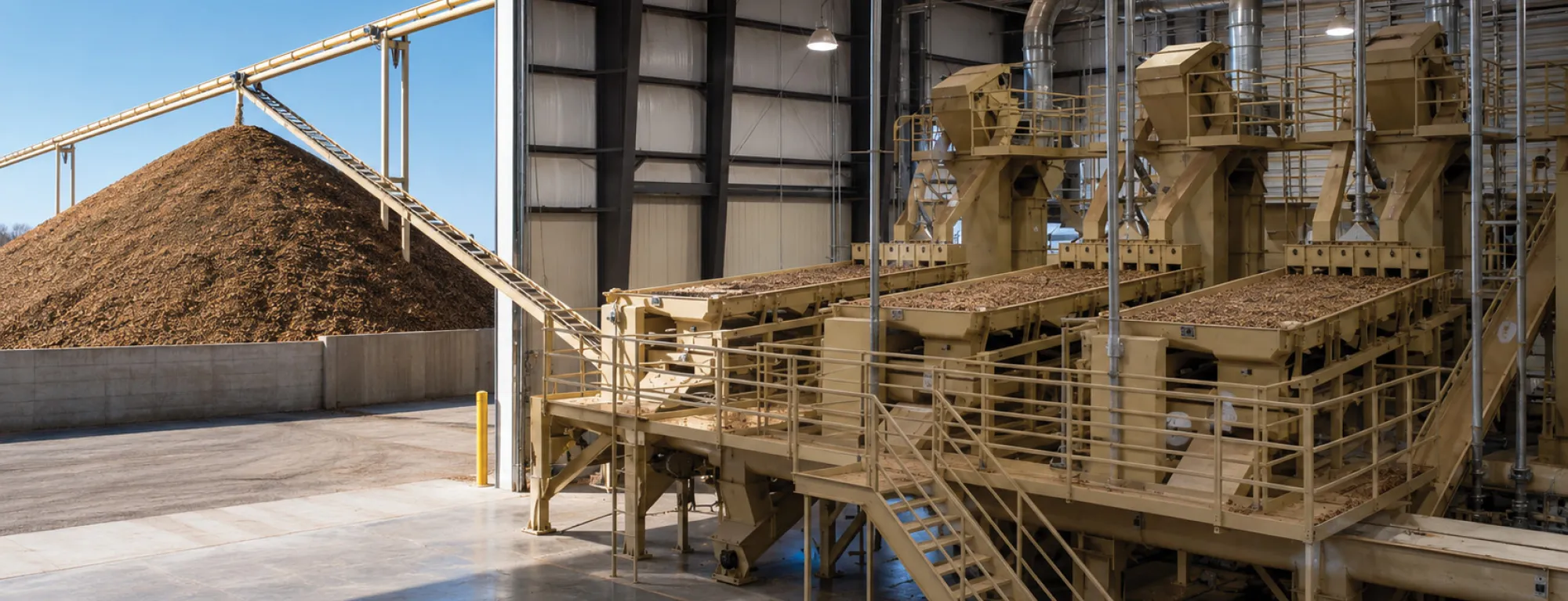 On left is a tall mound of almond hulls at the end of a conveyer running from an almond processing plant. On right are three machines, side by side, with trays full of nuts