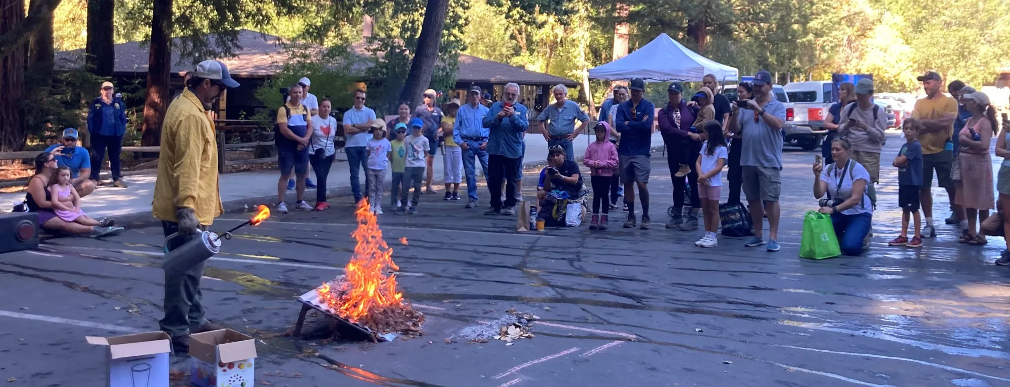A crowd of kids and adults watch a fire expert light a fire on an inclined board 