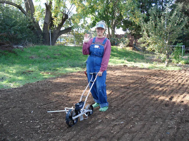 A person in denim overalls uses an Earth Way precision seeder in a tilled garden, surrounded by trees and grassy areas.