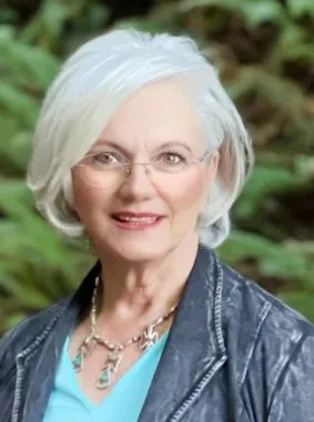 Sherida Phibbs has white hair and glasses and is wearing a blue shirt and charcoal jacket. 