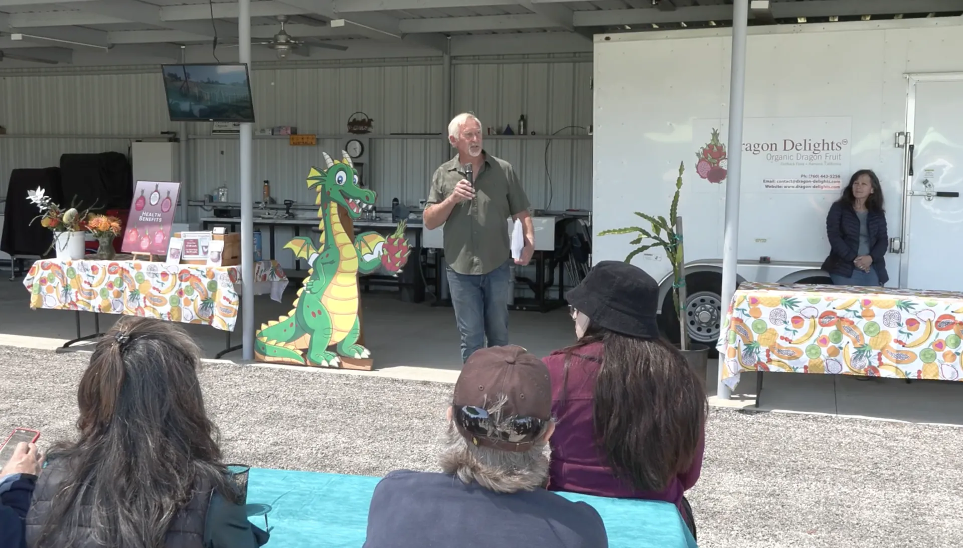 White haired man speaks into mic as three people in front of the look on. Behind him is cardboard cutout of a cartoon dragon holding a red dragonfruit.