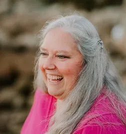 P Pearson smiles while wearing a pink blouse. She has long gray hair. 