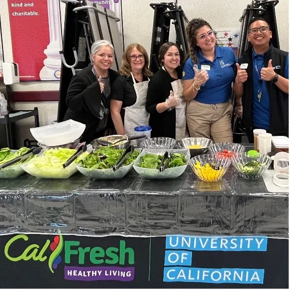 Sue Lafferty and 4 other people stand behind a long table displaying serving dishes of lettuce and other salad ingredients.