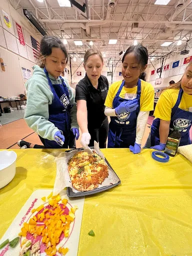 Three young children are being helped by an adult to make a pizza