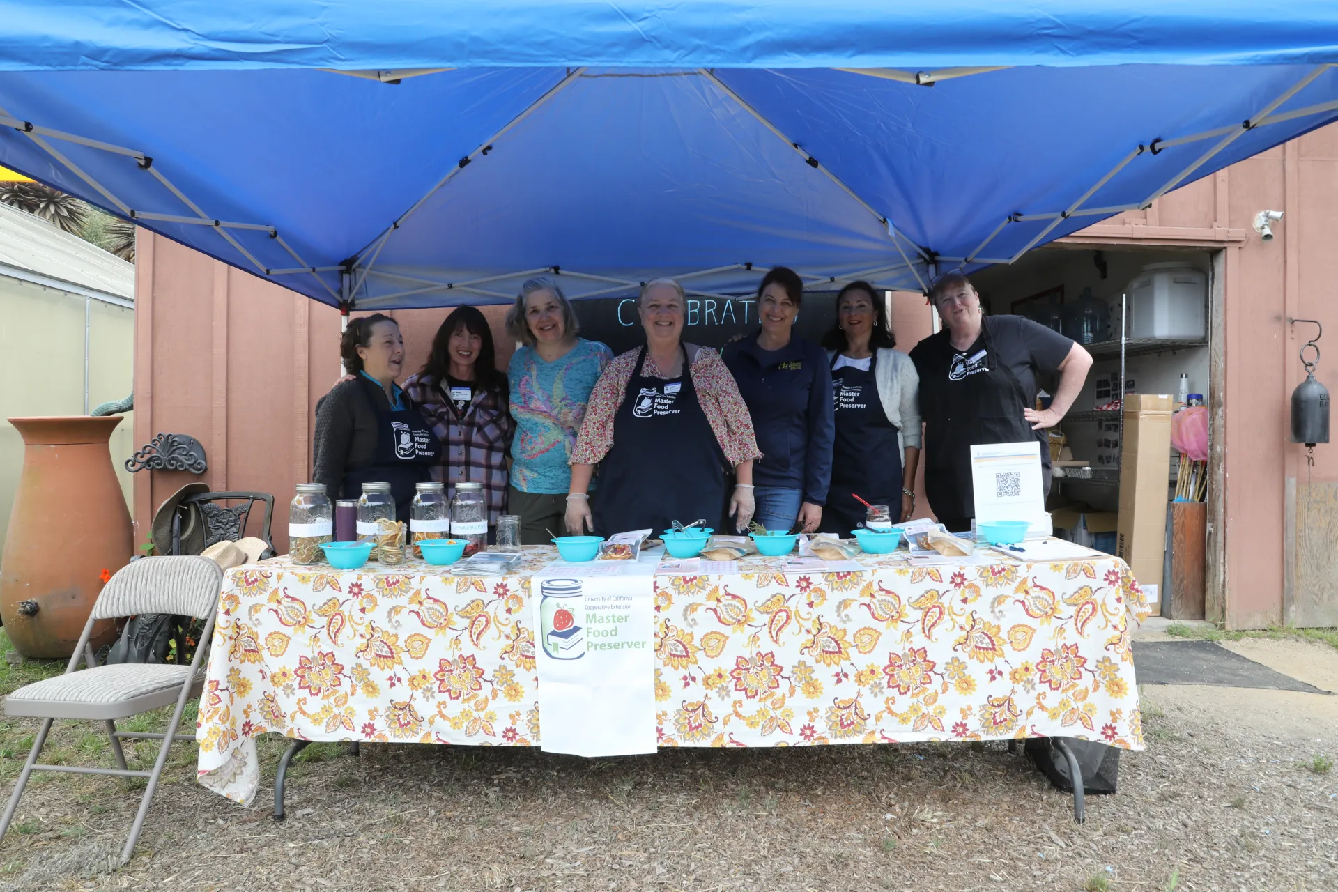 Seven women pose under a tent, behind a table with bowls and a sign that reads, "Master Food Preserver."