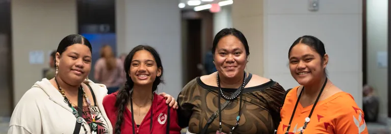 Four brown-skinned femmes wearing Indigenous prints and Ignite badges smile with their arms around one another.
