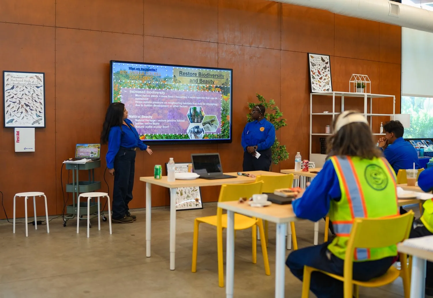 Two Long Beach Conservation Corps Members dressed in a blue top and black bottom uniform present their stewardship project to a room full of their peers using a projector and powerpoint slide set as they discuss the importance of polinators