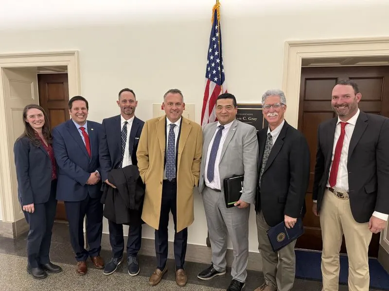 A woman and six men wearing suits stand in front of U.S. flag and Rep. Adam Gray's office plaque.
