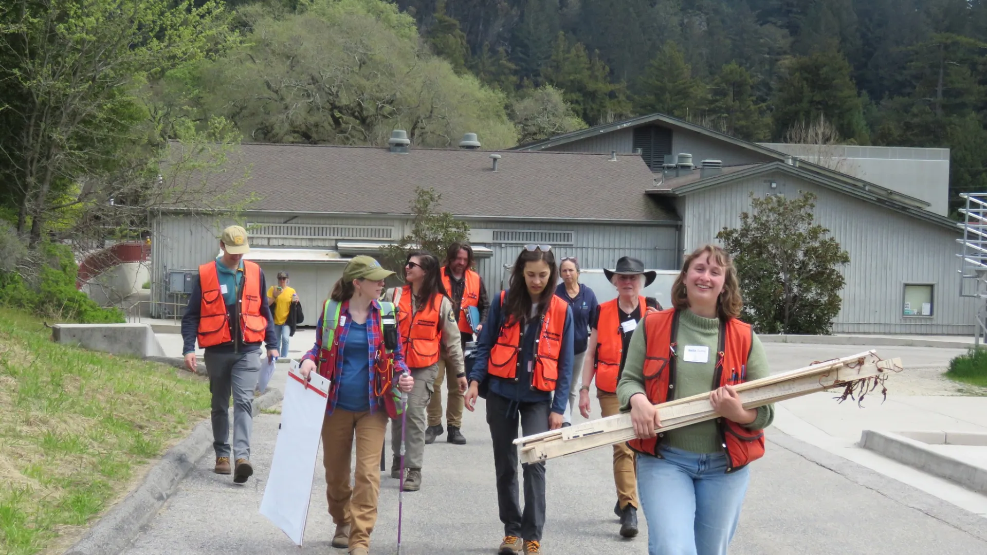 A group of instructors, walking in loose rows, holds wood pieces, an easel and papers as they walk away from a gray building and head into the woods for Tree School