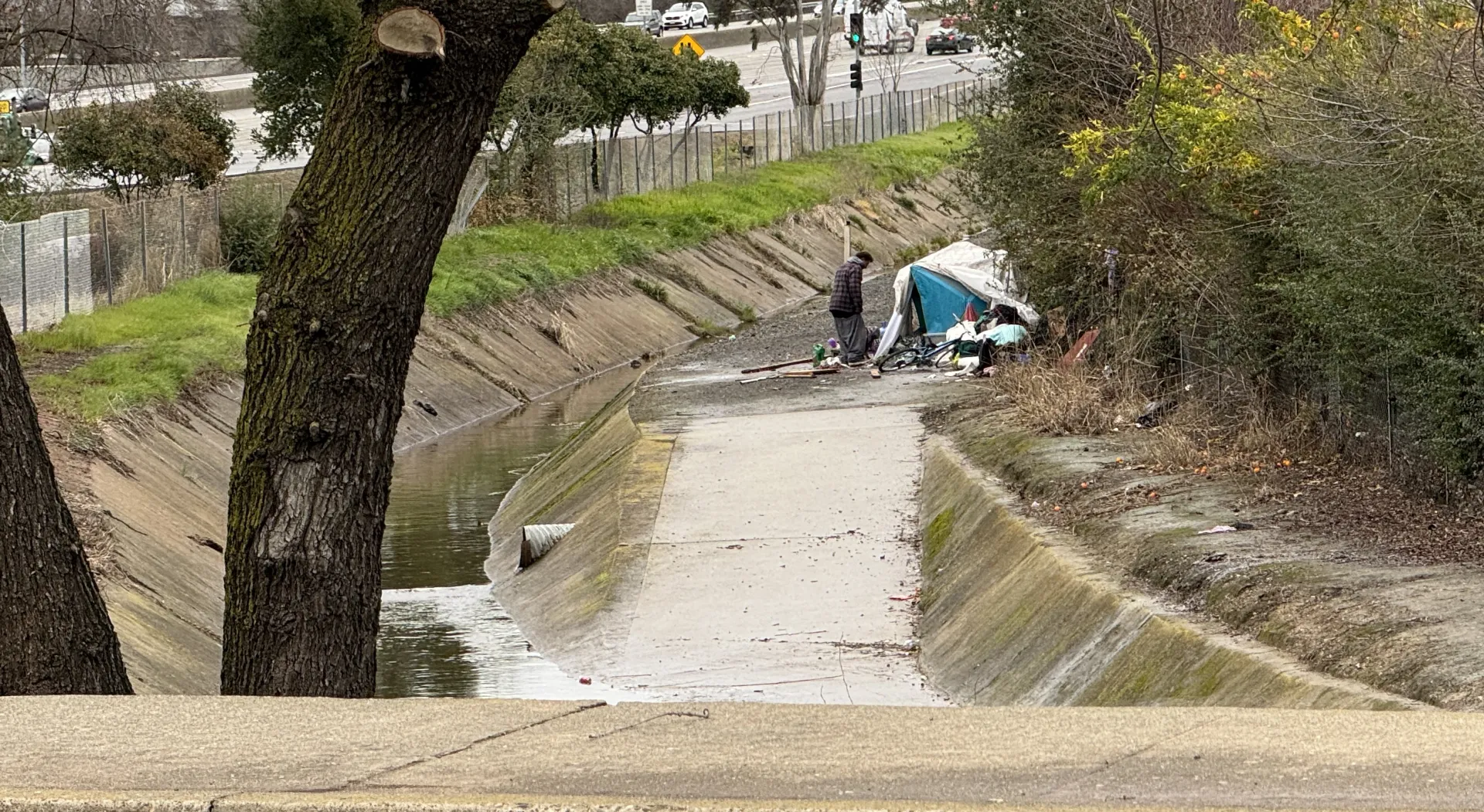 A man stands outside a tent located on the banks of a canal that has a low level of water