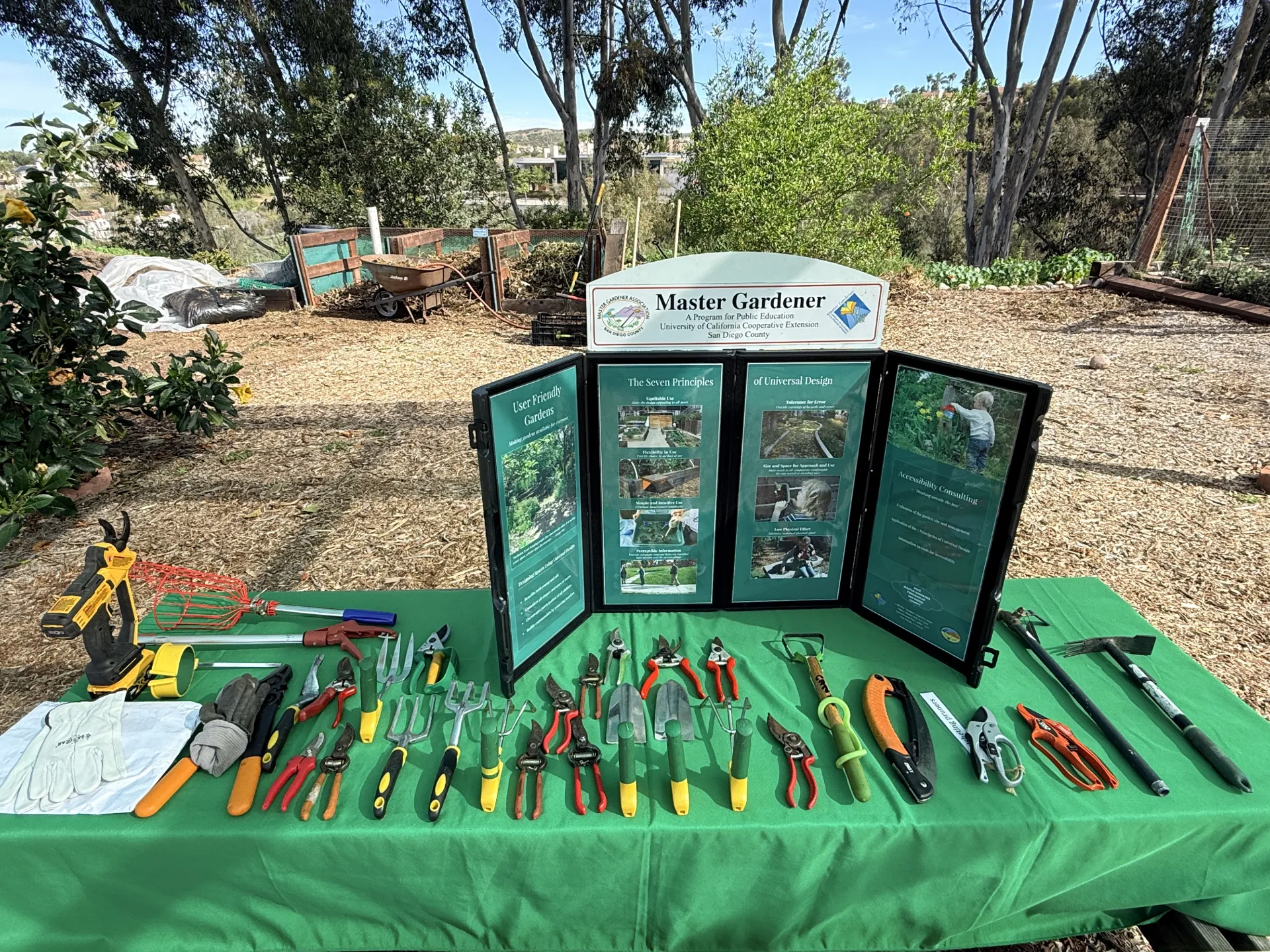 A display of gardening tools on a green tablecloth in front of a Master Gardener informational tri-fold board. The garden tools are specialized for adaptive uses. In the background is a garden area with bark paths and compost bins.
