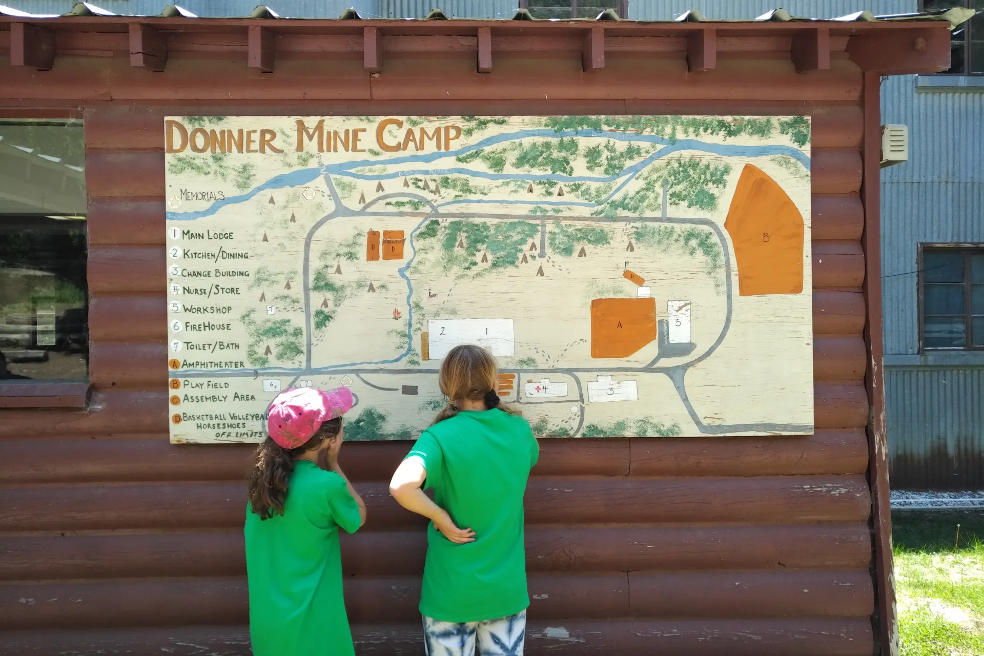Two young campers are standing in front of a large hand-drawn map of the Donner Mine Camp and looking at the map. The map is attached to the front of the main camp building.