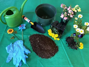 On a green tablecloth are many items including blue gloves, silk flowers, a green plastic watering can, a hand trowel, a dark green pot, a small pile of dark soil, and three plastic pot packs of yellow, pink and white flowers.